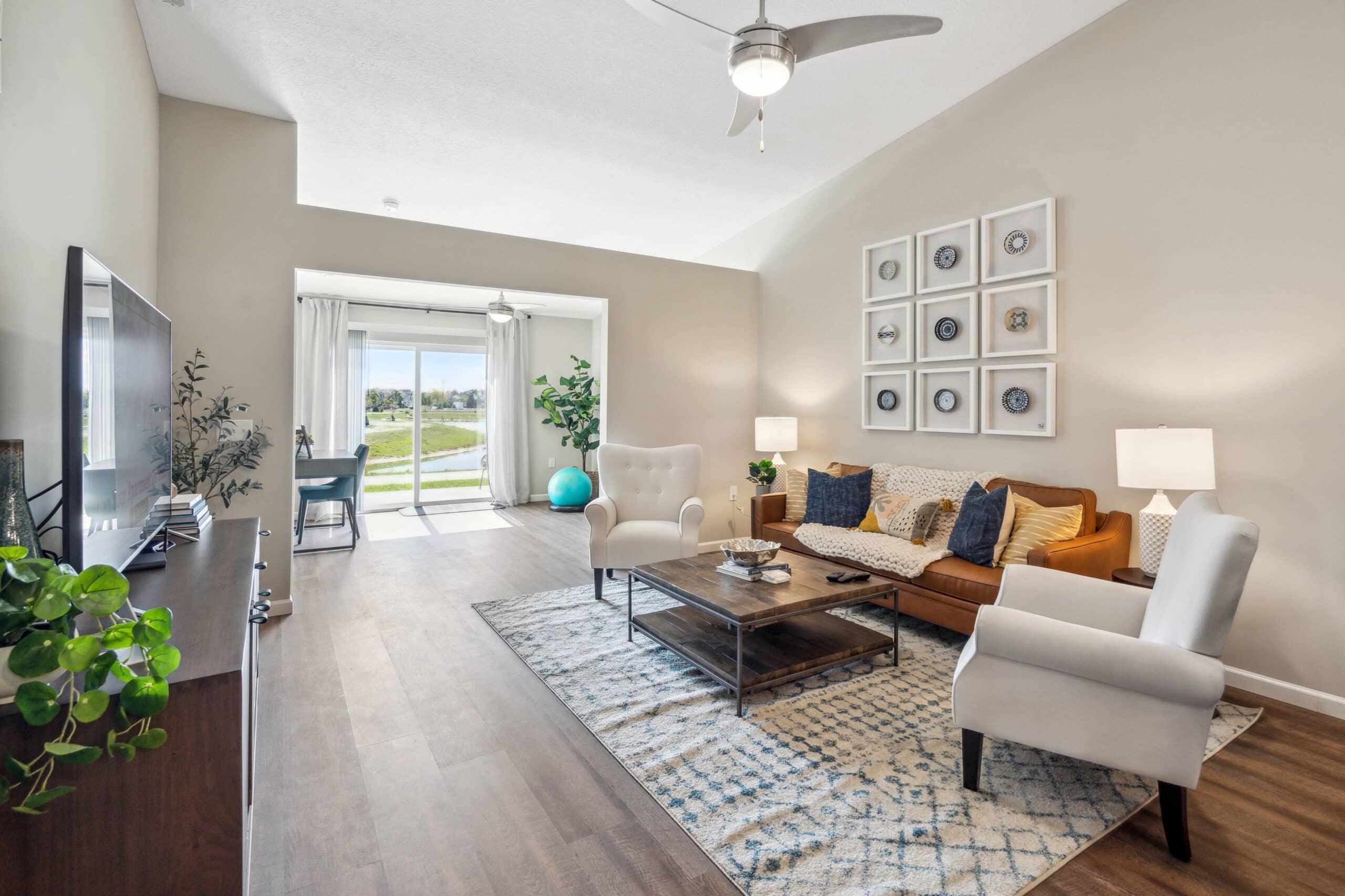 A modern living room with a couch, two armchairs, a coffee table, and a TV. Large windows show a scenic view outside. Decor includes plants, lamps, and wall art. Hardwood flooring is visible.