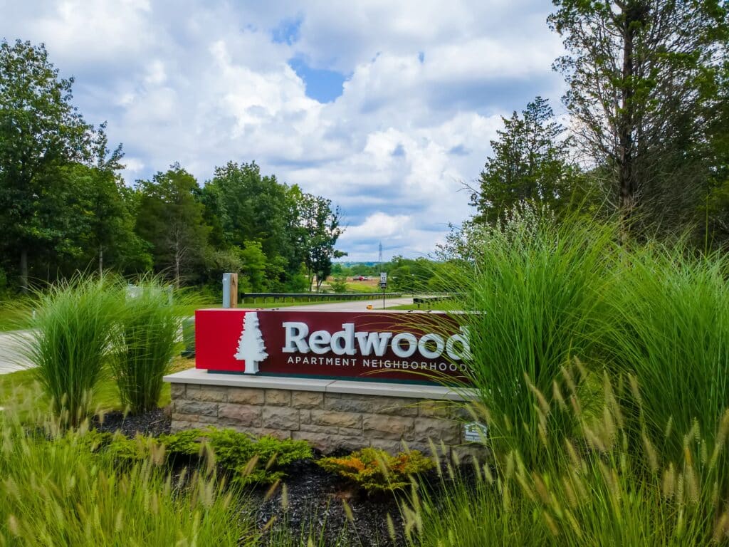A stone and red sign reads “Redwood Apartment Neighborhoods,” surrounded by tall grass and trees under a partly cloudy sky—tour Redwood Vandalia Apartments in Dayton and experience this inviting setting.