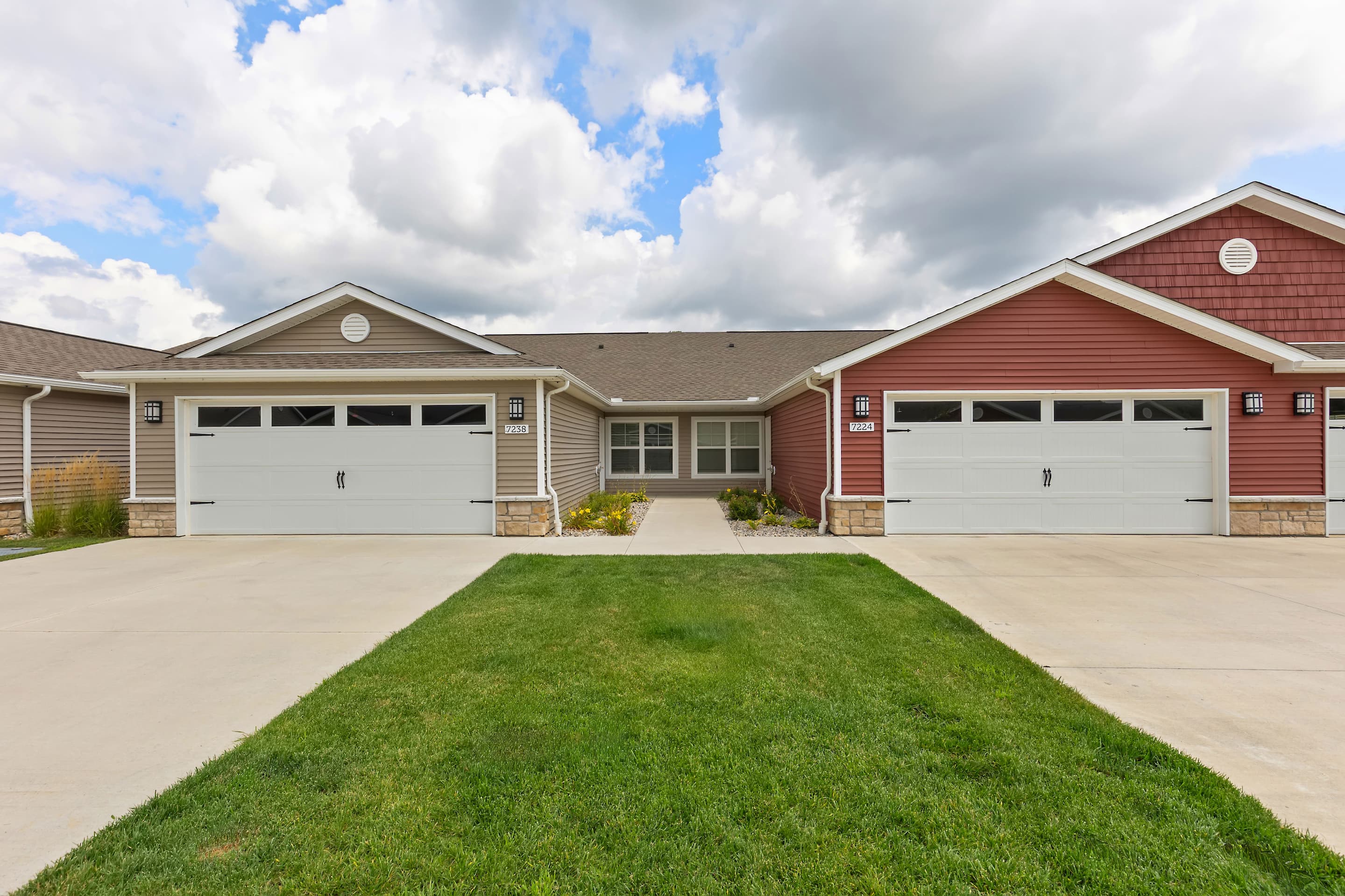 Tour Redwood Vandalia Apartments in Dayton, featuring two connected single-story homes with attached double garages, beige and red siding, and a shared walkway leading to the main entrances.