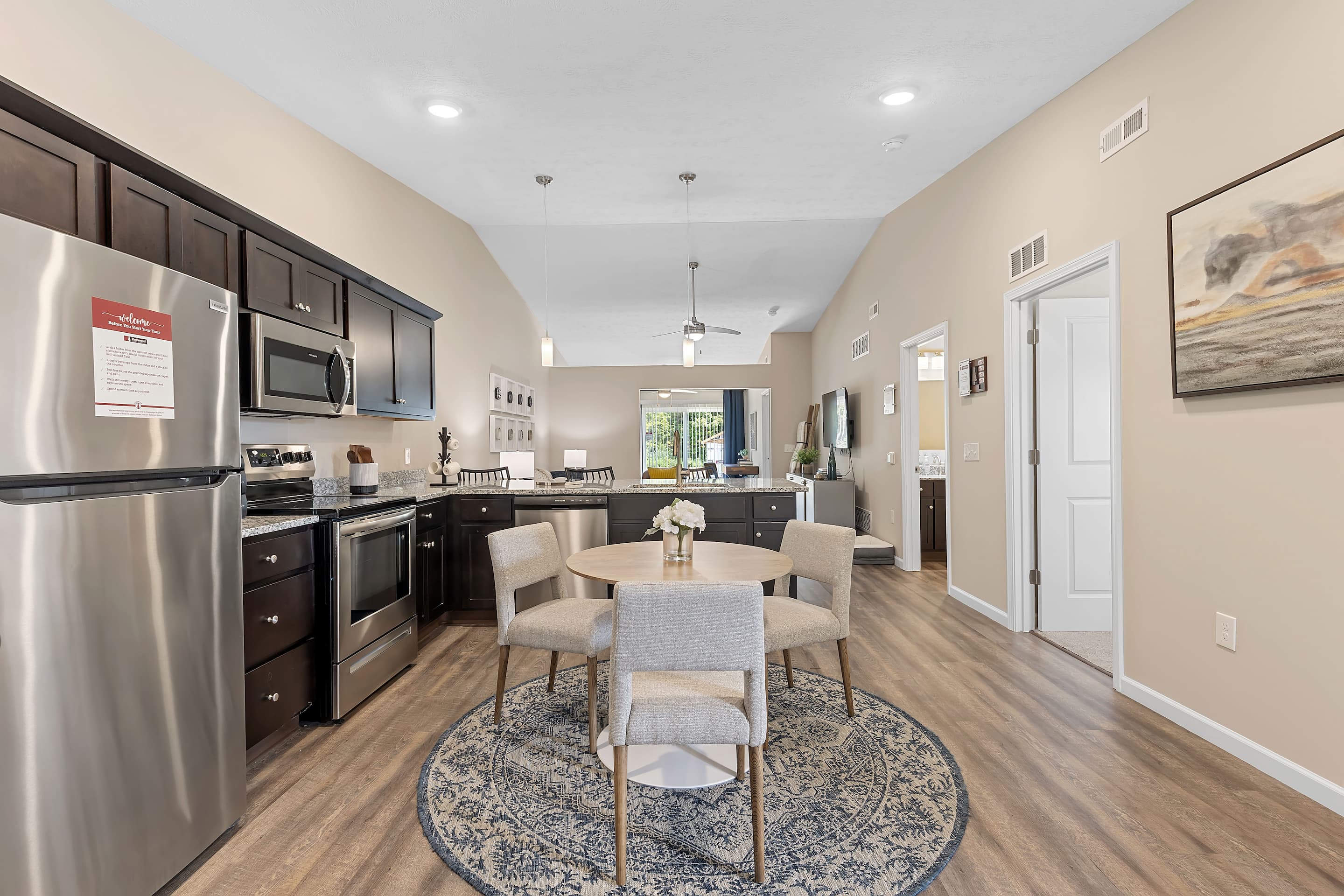 Tour Redwood Vandalia Apartments in Dayton and discover a modern kitchen and dining area with stainless steel appliances, dark cabinets, a round table on a patterned rug, and a large window in the background.