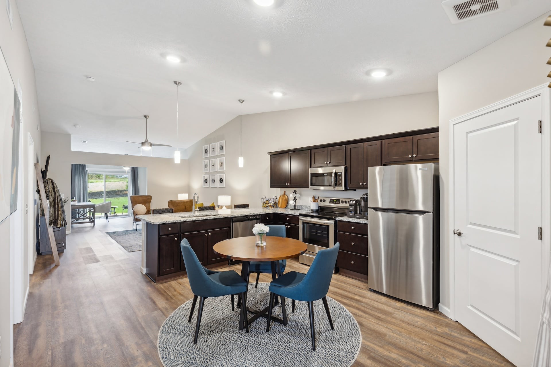 Modern kitchen and dining area with dark wood cabinets, stainless steel appliances, a round table with two blue chairs on a patterned rug, and a view into a living room with sliding doors to a patio.