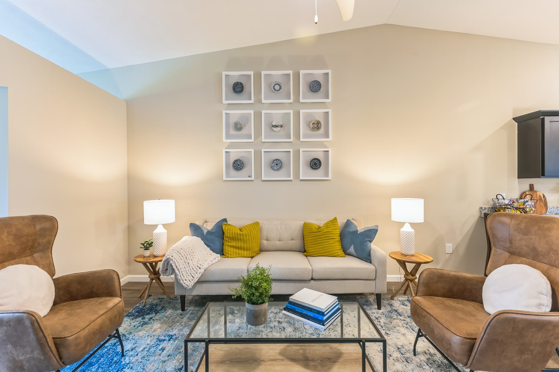 A modern living room with a beige sofa, yellow pillows, two brown armchairs, a rectangular coffee table, and framed art on the wall. Two table lamps are on either side of the sofa.