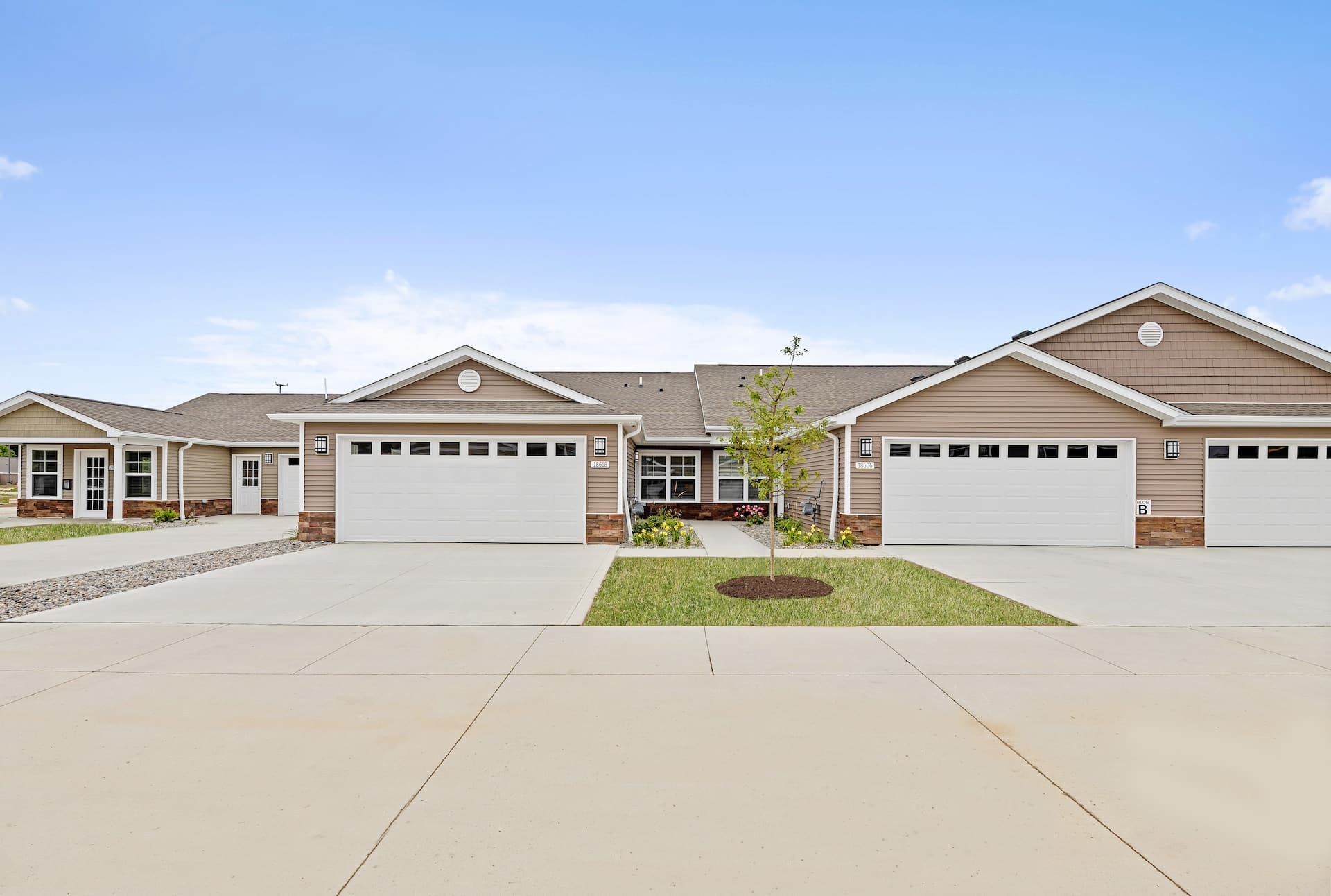 A row of connected beige one-story houses with two-car garages and driveways under a clear blue sky.