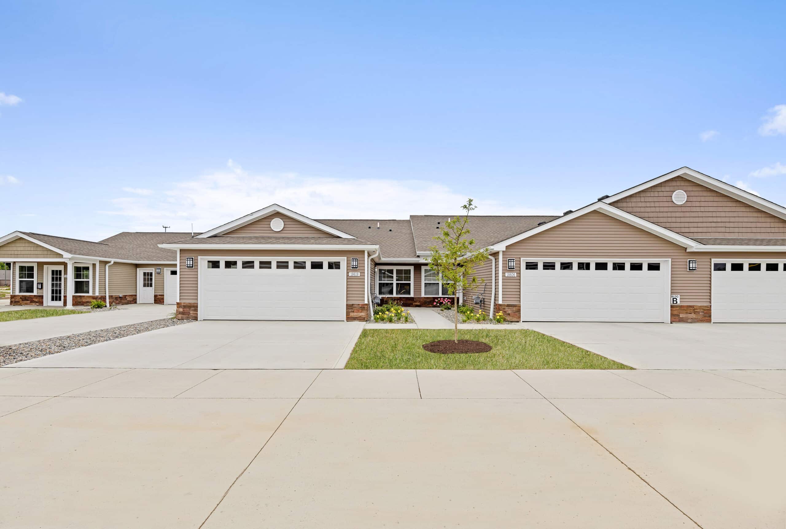 A row of attached single-story houses with beige siding and white garage doors, each featuring driveways and small patches of grass, under a clear blue sky.