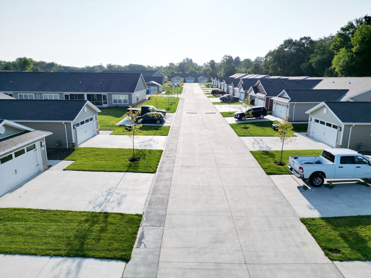 Aerial view of a suburban neighborhood with a straight road, lined with single-story houses, driveways, and neatly maintained lawns. Several vehicles are parked along the street.