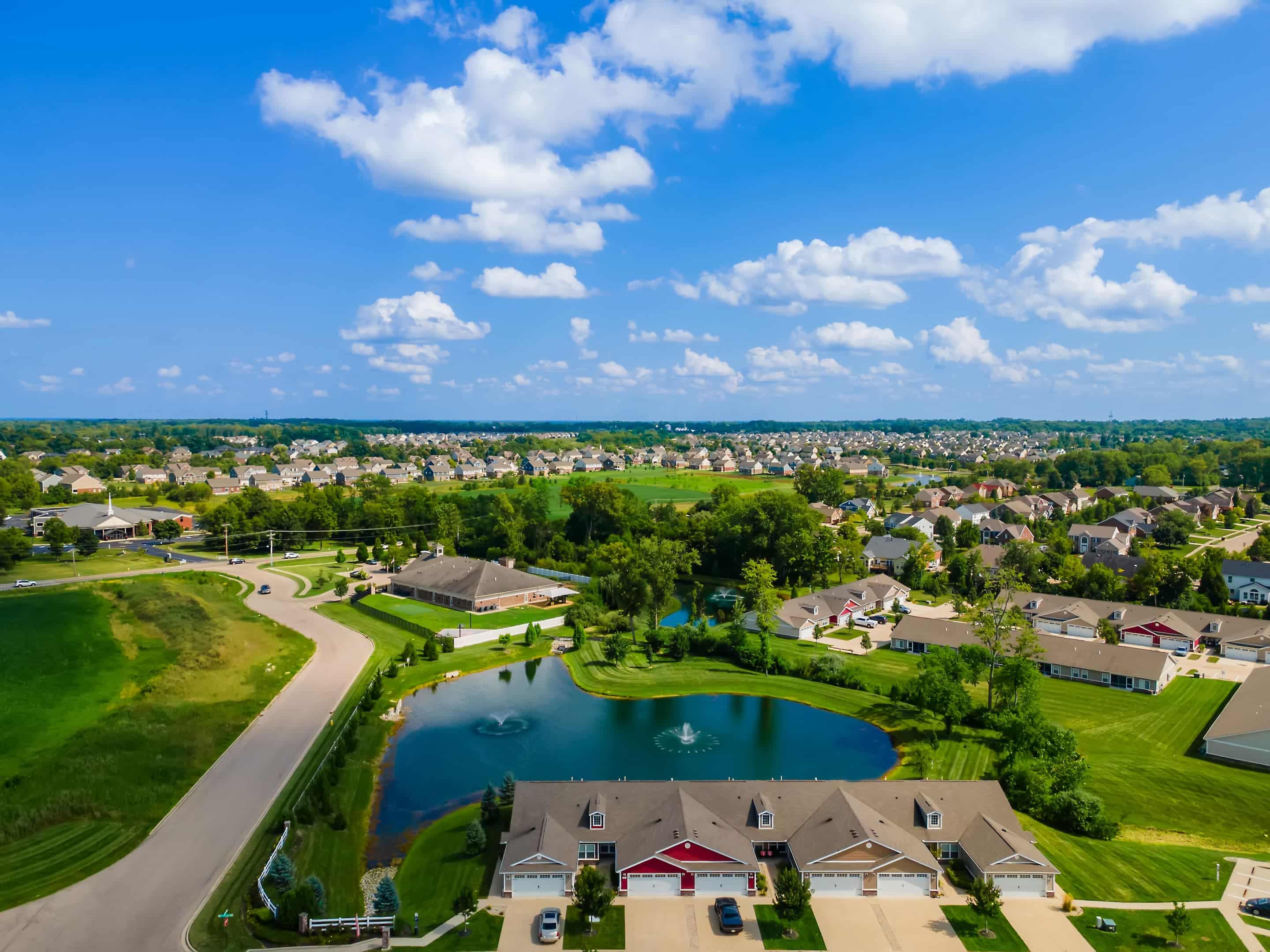 Aerial view of a suburban neighborhood with houses, a small pond with fountains, green lawns, and roads under a partly cloudy sky.