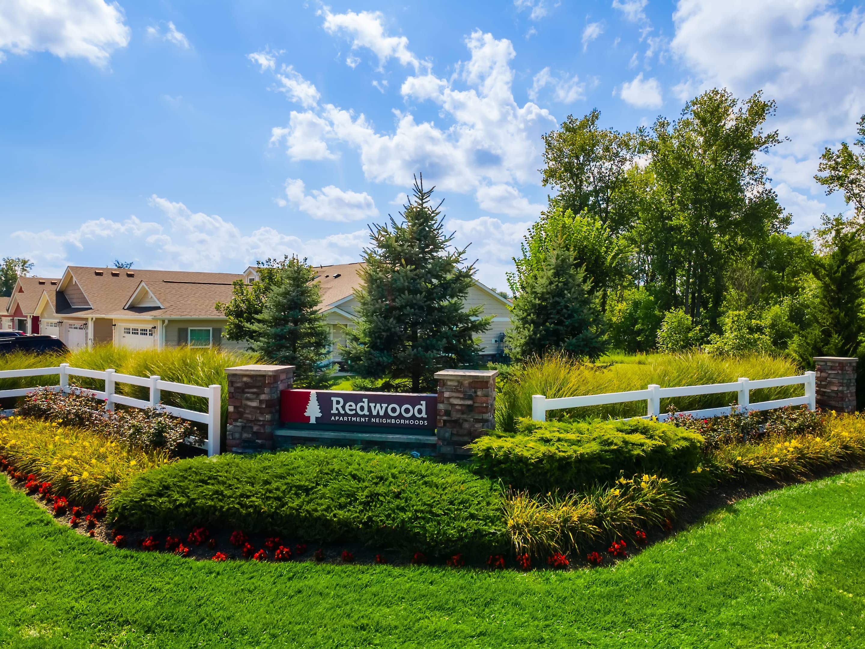 A landscaped entrance with a "Redwood Apartment Neighborhoods" sign, white fence, shrubs, and houses in the background under a partly cloudy sky.
