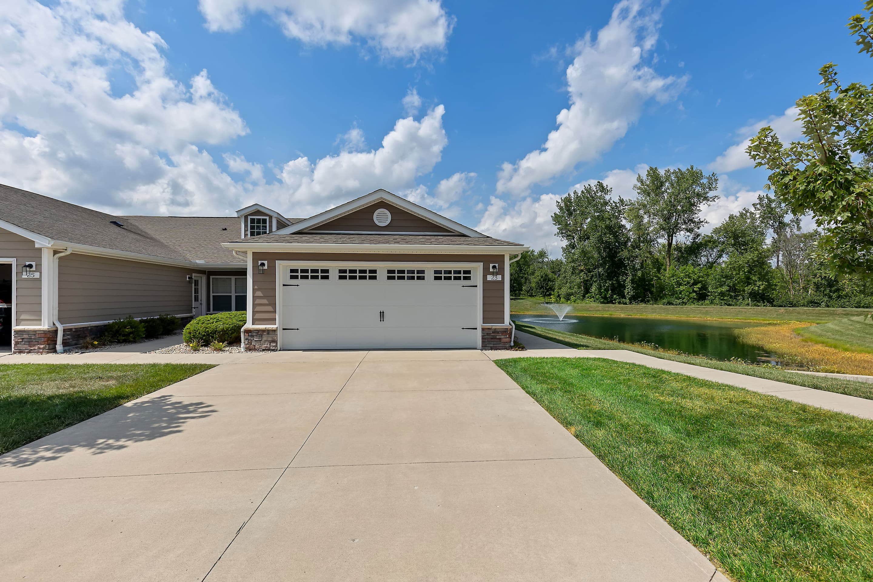 A beige suburban house with a double garage, concrete driveway, green lawn, and pond with a fountain in the background on a sunny day.