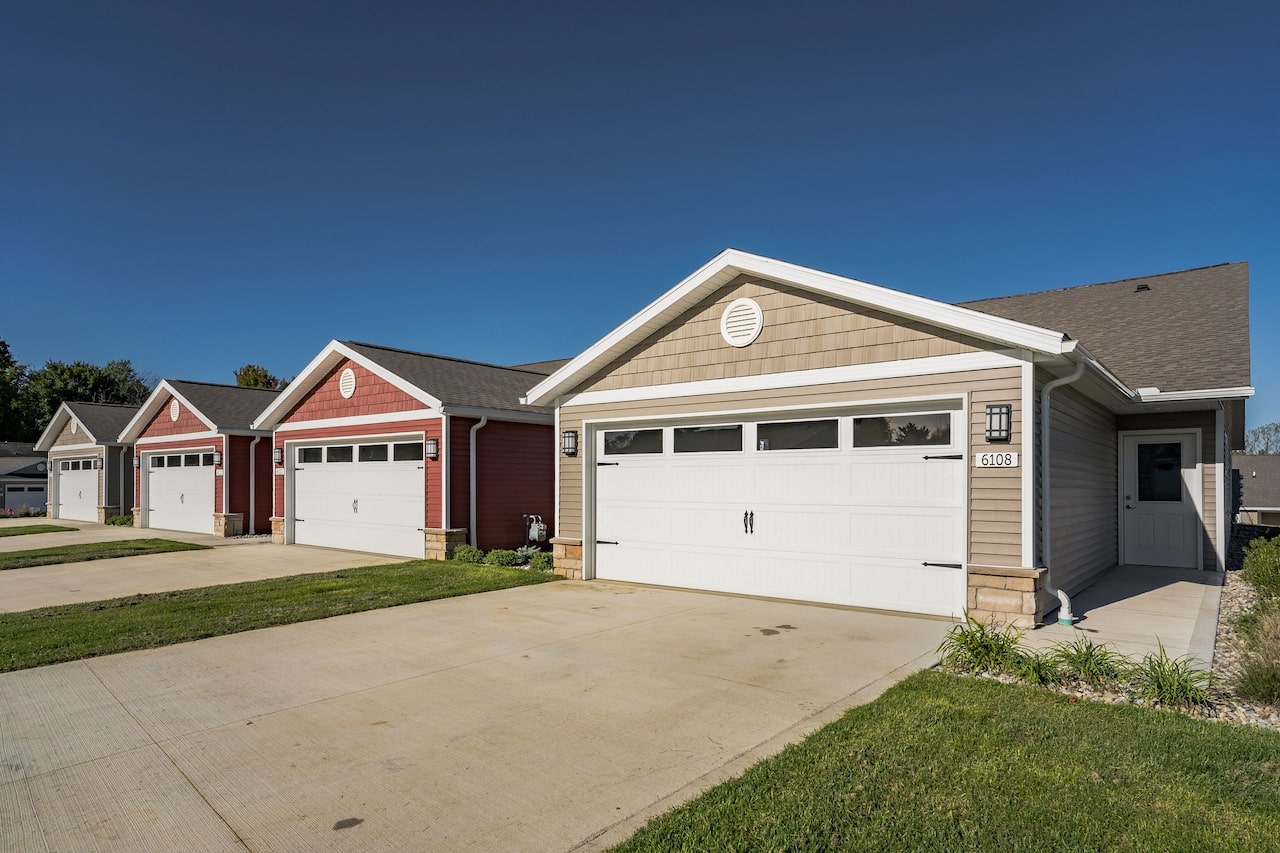 A row of single-story homes with garages, featuring different colored facades, in a suburban neighborhood under a clear blue sky.