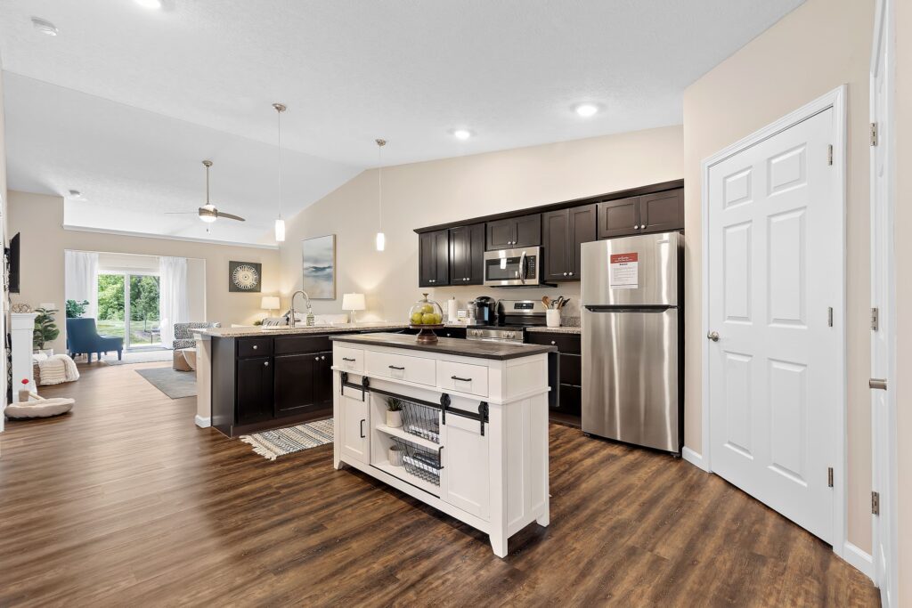 Modern kitchen with dark cabinets, stainless steel appliances, white island, and wood flooring; open layout leads to dining and living areas with glass doors to outside.