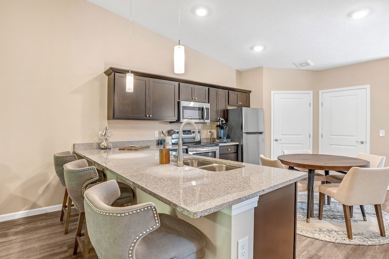 Modern kitchen with granite countertops, bar stools, dark wood cabinets, and stainless steel appliances. Adjacent dining area with a round table and beige chairs on a rug.