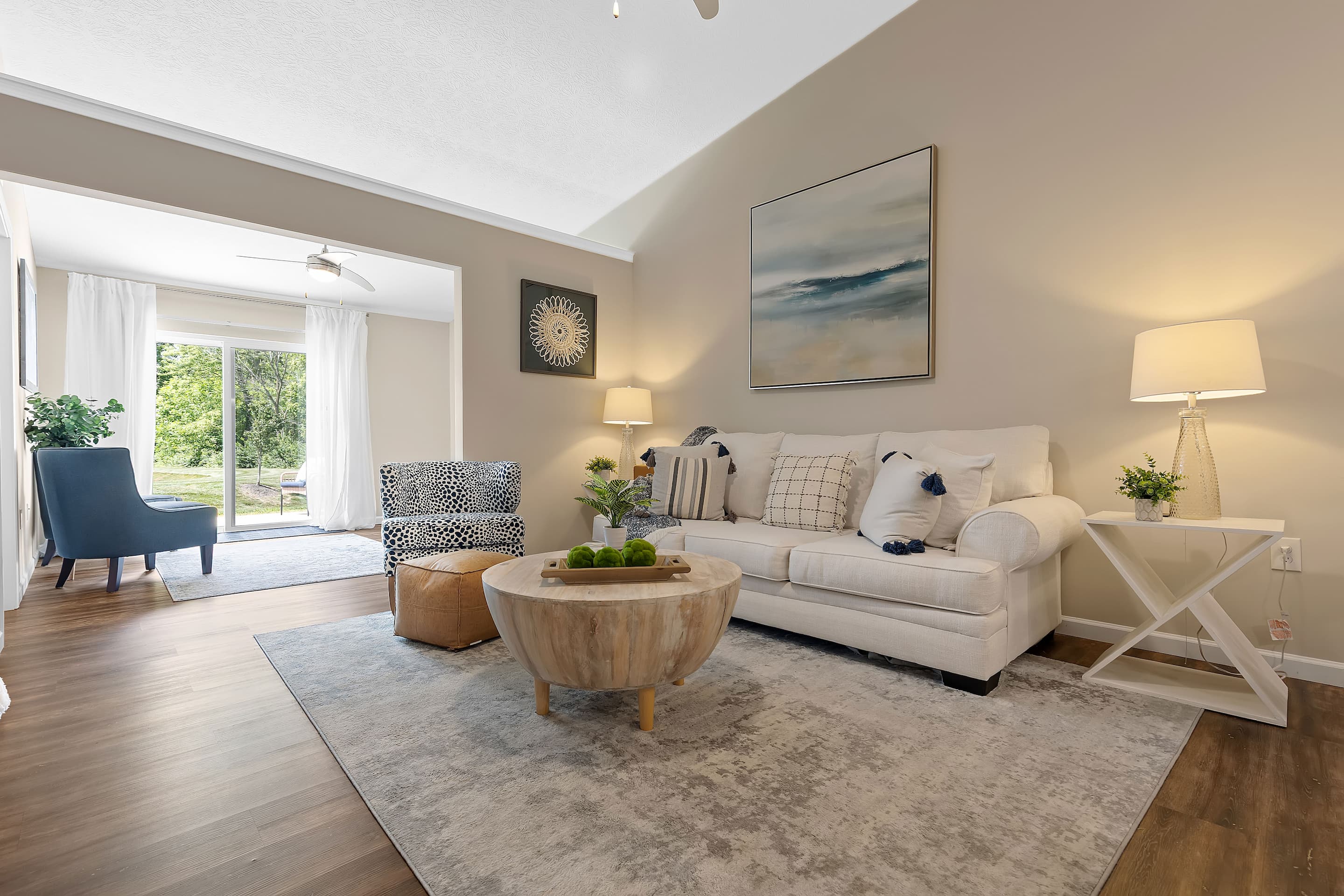A modern living room with a white sofa, round wooden coffee table, two chairs, lamps, wall art, a rug, and large windows letting in natural light.