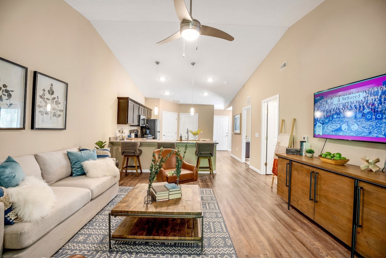 Modern living room with beige walls, a ceiling fan, and wood flooring. A kitchen area is in the background. A sofa, coffee table, and TV are visible along with decorative items and artwork.