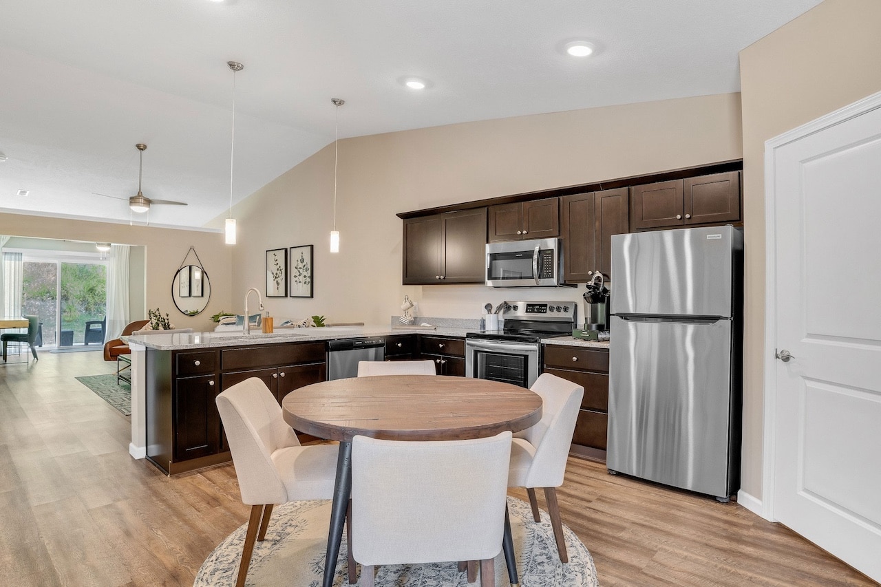 A modern kitchen with dark wood cabinets, stainless steel appliances, and a round dining table with four chairs. An open living area is visible in the background.