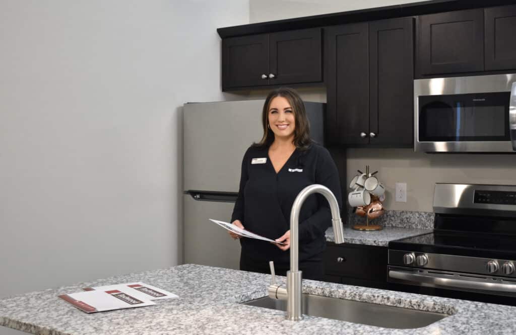 A woman stands in a modern kitchen with granite countertops, holding papers. A stainless steel refrigerator and stove are in the background.