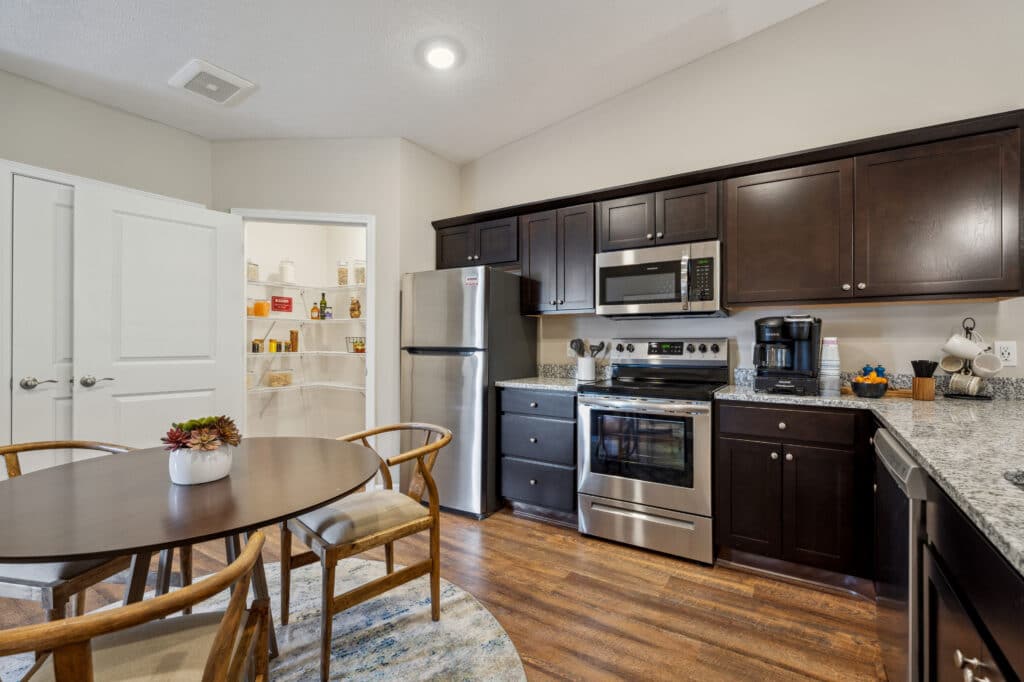 Modern kitchen with dark wood cabinets, stainless steel appliances, and granite countertops. A round dining table with chairs is in the foreground. Open pantry door visible in the back.