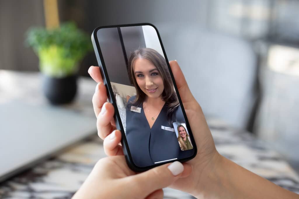 A person holds a smartphone showing a video call with two women on screen. One is the main focus, and the other appears in a smaller window. A blurred plant is in the background.