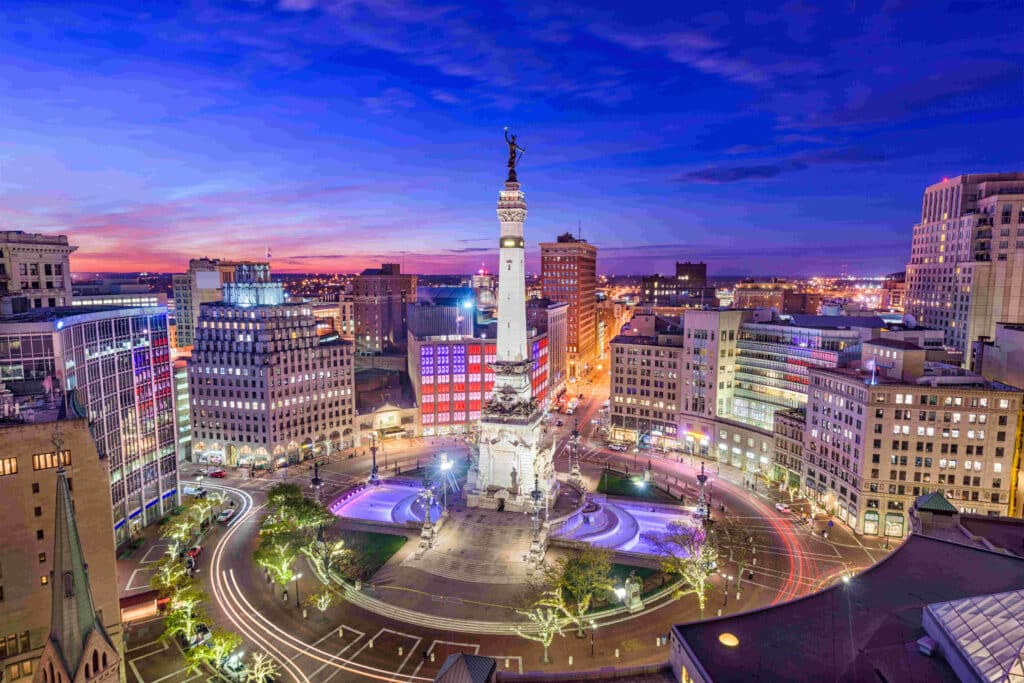Aerial view of a city plaza at dusk, with a central monument surrounded by traffic. Buildings are illuminated, and light trails from vehicles are visible around the monument.