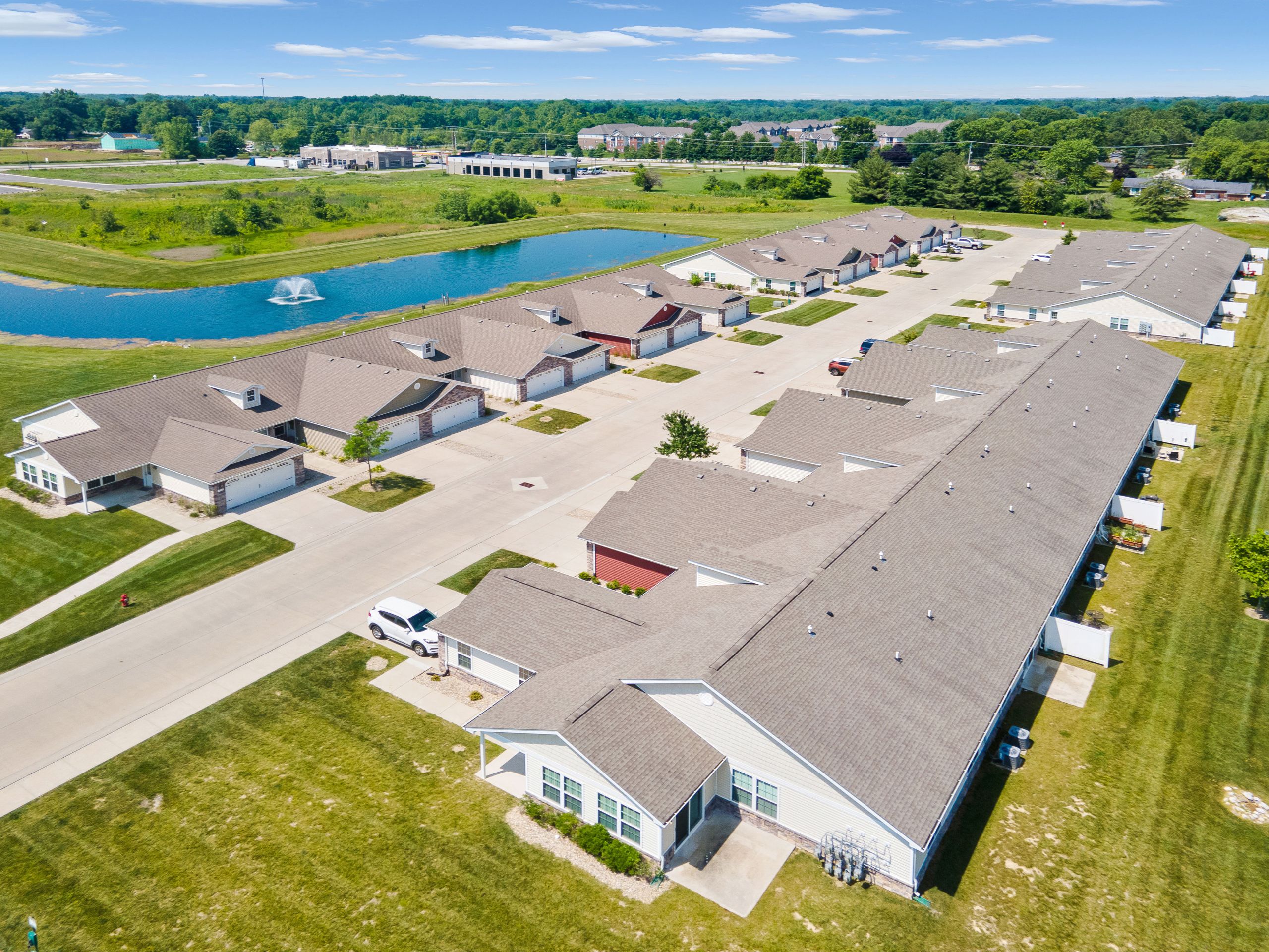 Aerial view of a suburban residential community with rows of single-story homes, driveways, green lawns, and a pond with a fountain.