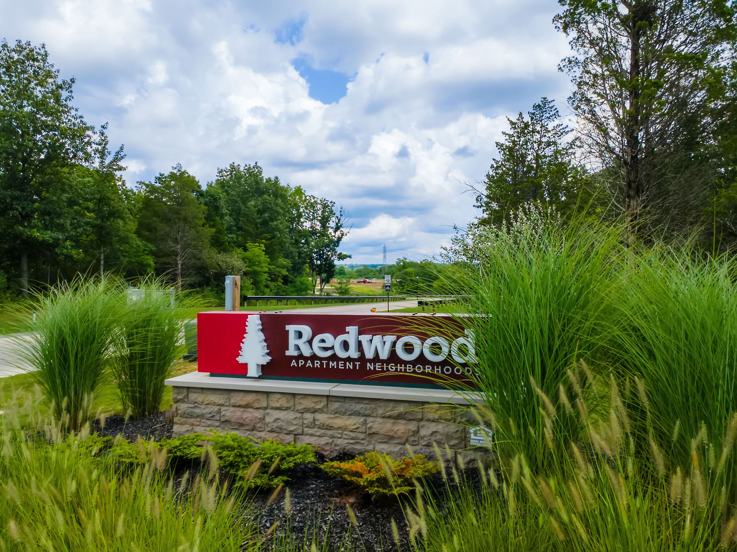 A sign reading "Redwood Apartment Neighborhood" stands among tall green grass and trees under a partly cloudy sky.