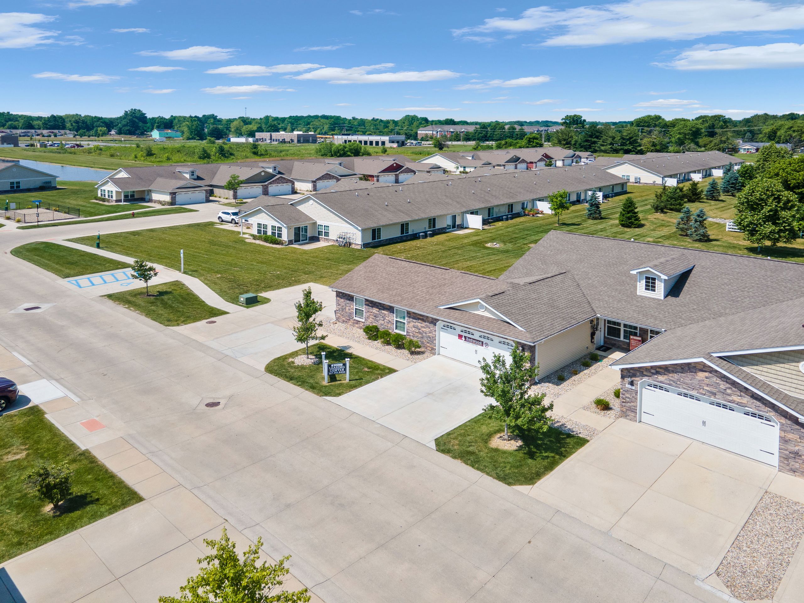 Aerial view of a suburban neighborhood with single-story houses, green lawns, sidewalks, and a nearby pond under a blue sky.