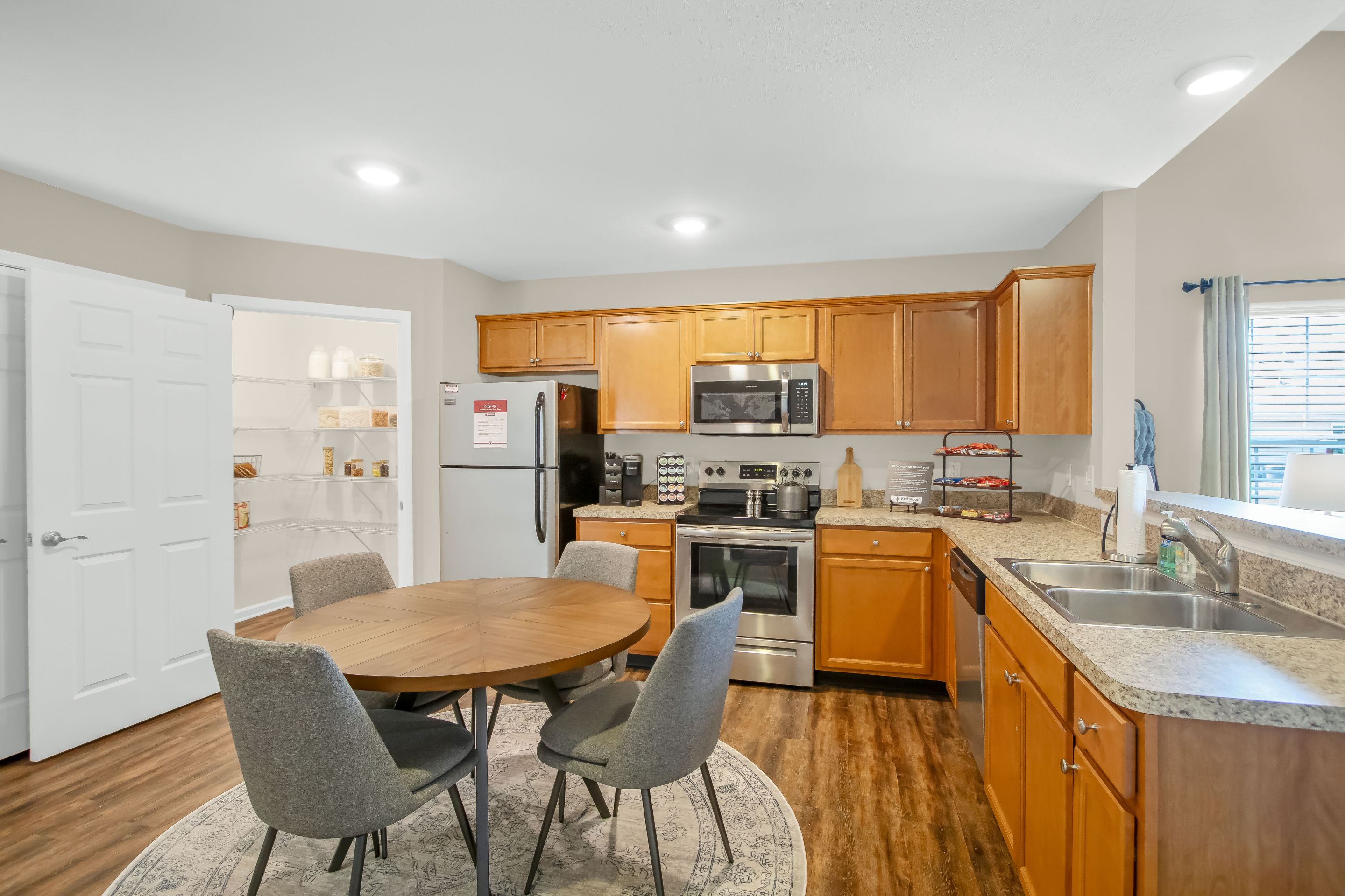 A modern kitchen with wooden cabinets, stainless steel appliances, a round dining table with four gray chairs, and an open pantry with shelves.