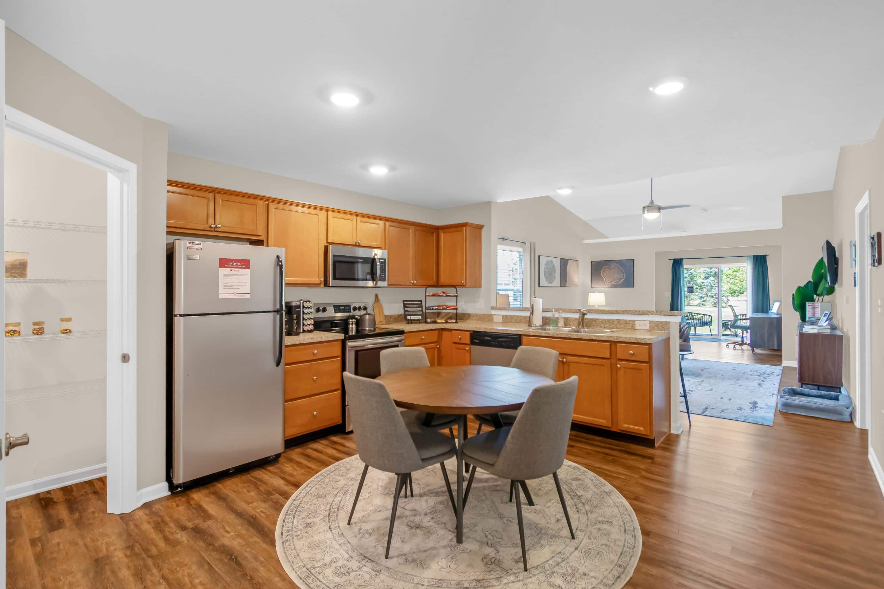 Modern kitchen with stainless steel appliances, wooden cabinets, a round dining table with four chairs on a rug, and an open view into the living and dining area.