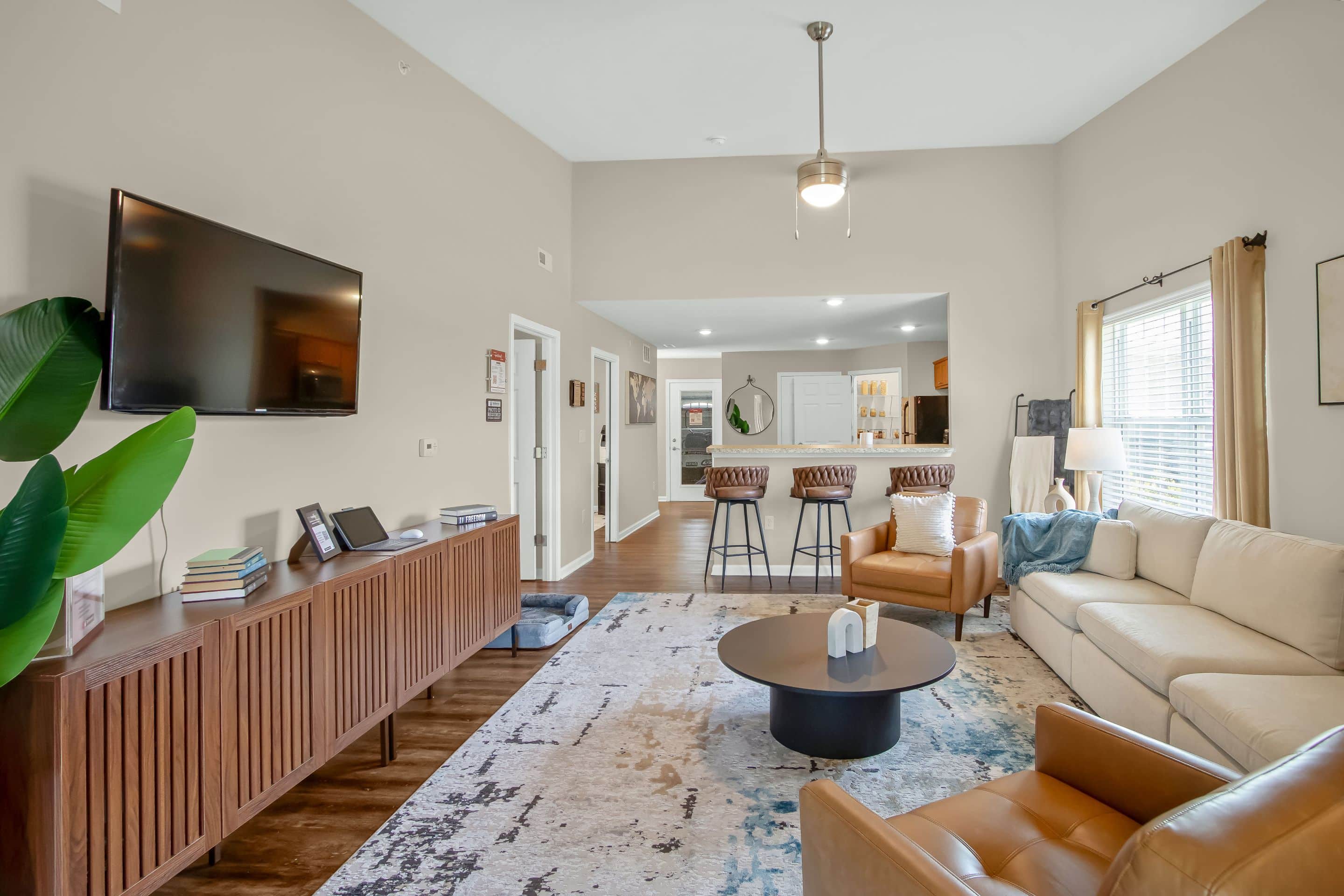 A modern living room with a TV mounted on the wall, a wooden console, a beige sofa, a brown armchair, a round coffee table, and a breakfast bar with stools facing the kitchen.