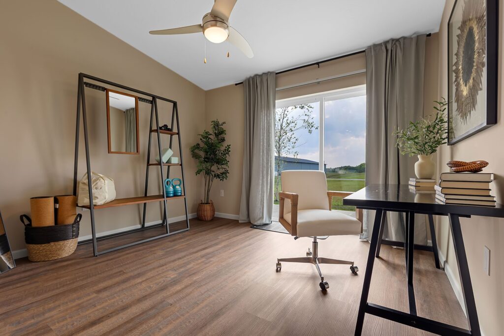 Home office with wooden floor, black desk, white chair, open shelving unit, potted plants, neutral walls, and large window with curtains overlooking a grassy outdoor view.