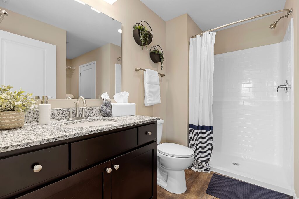 Modern bathroom with a granite countertop, dark wood vanity, white toilet, walk-in shower with striped curtain, and two potted plants on the wall.