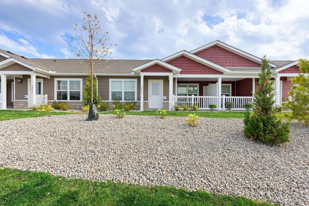 Single-story townhouse with a porch, large windows, and a gravel front yard with young trees and shrubs under a partly cloudy sky.