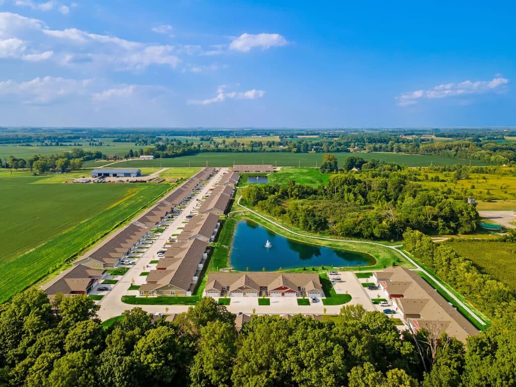 Aerial view of a suburban neighborhood with rows of houses, a central pond with a fountain, and surrounding fields and trees under a clear blue sky.