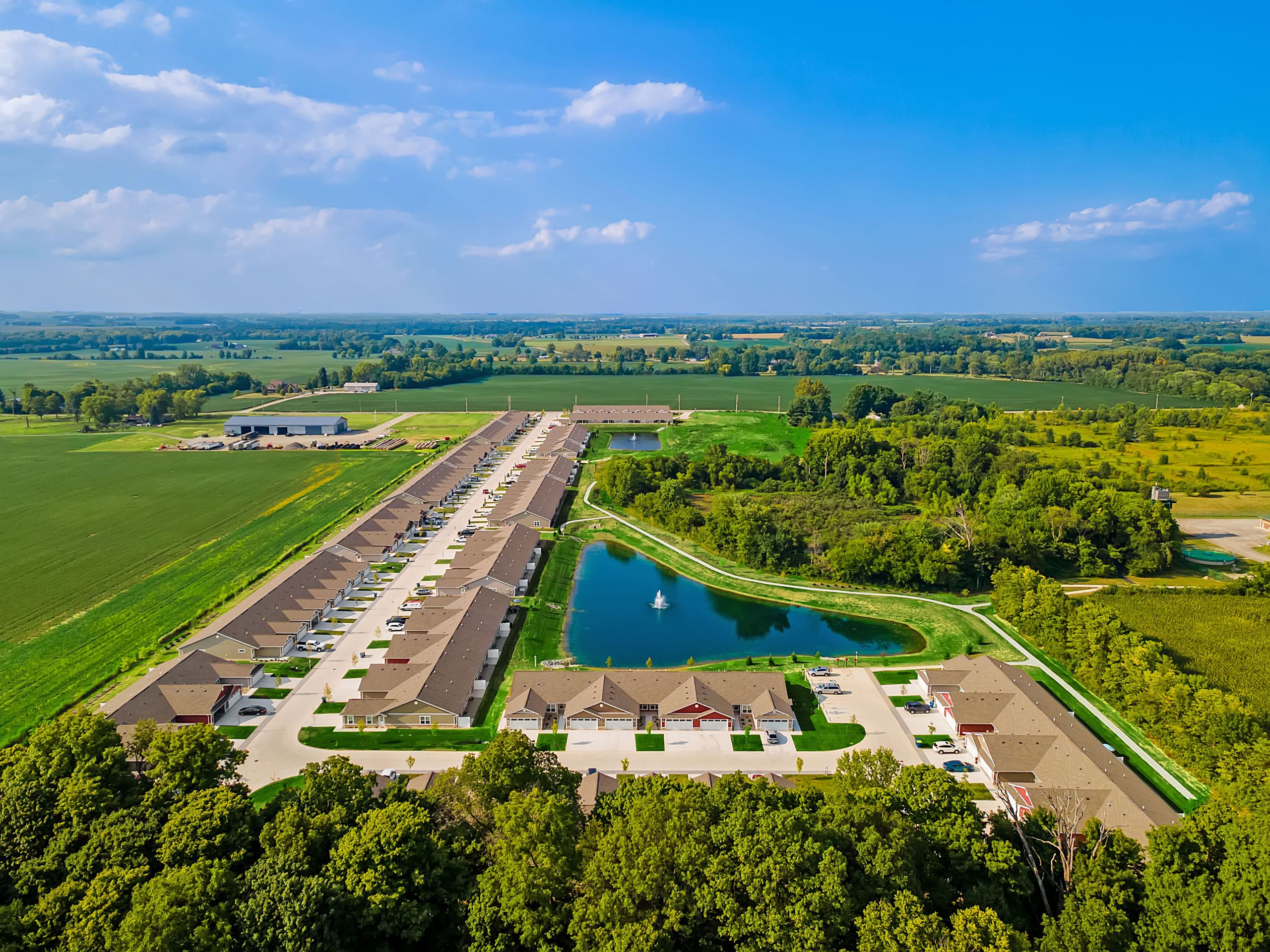 Aerial view of a suburban neighborhood with rows of houses, a central pond with a fountain, and surrounding fields and trees under a clear blue sky.