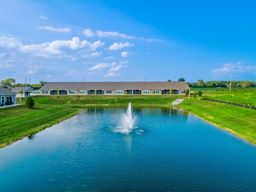 A small pond with a central water fountain is surrounded by green grass and a row of single-story buildings under a blue sky with scattered clouds.
