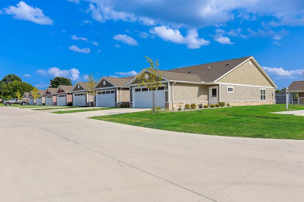 Row of single-story suburban houses with attached garages, green lawns, and a wide concrete street under a blue sky with scattered clouds.