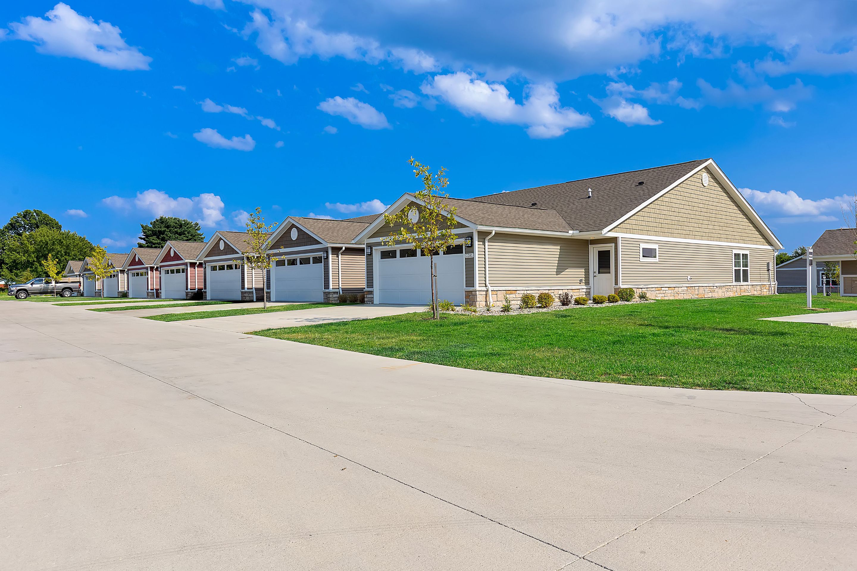 Row of single-story suburban houses with attached garages, green lawns, and a wide concrete street under a blue sky with scattered clouds.