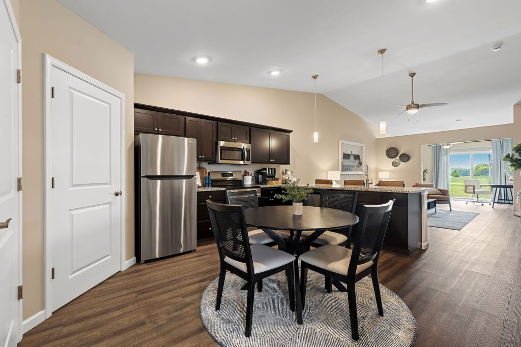 Modern kitchen and dining area with dark cabinets, stainless steel appliances, round table with four chairs, and a view into the living room and patio.