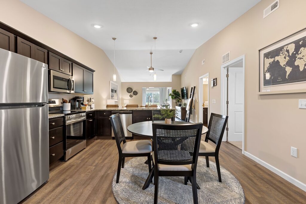 Modern kitchen and dining area with stainless steel appliances, dark cabinets, round dining table with four chairs, and a wall map, set on wood flooring.