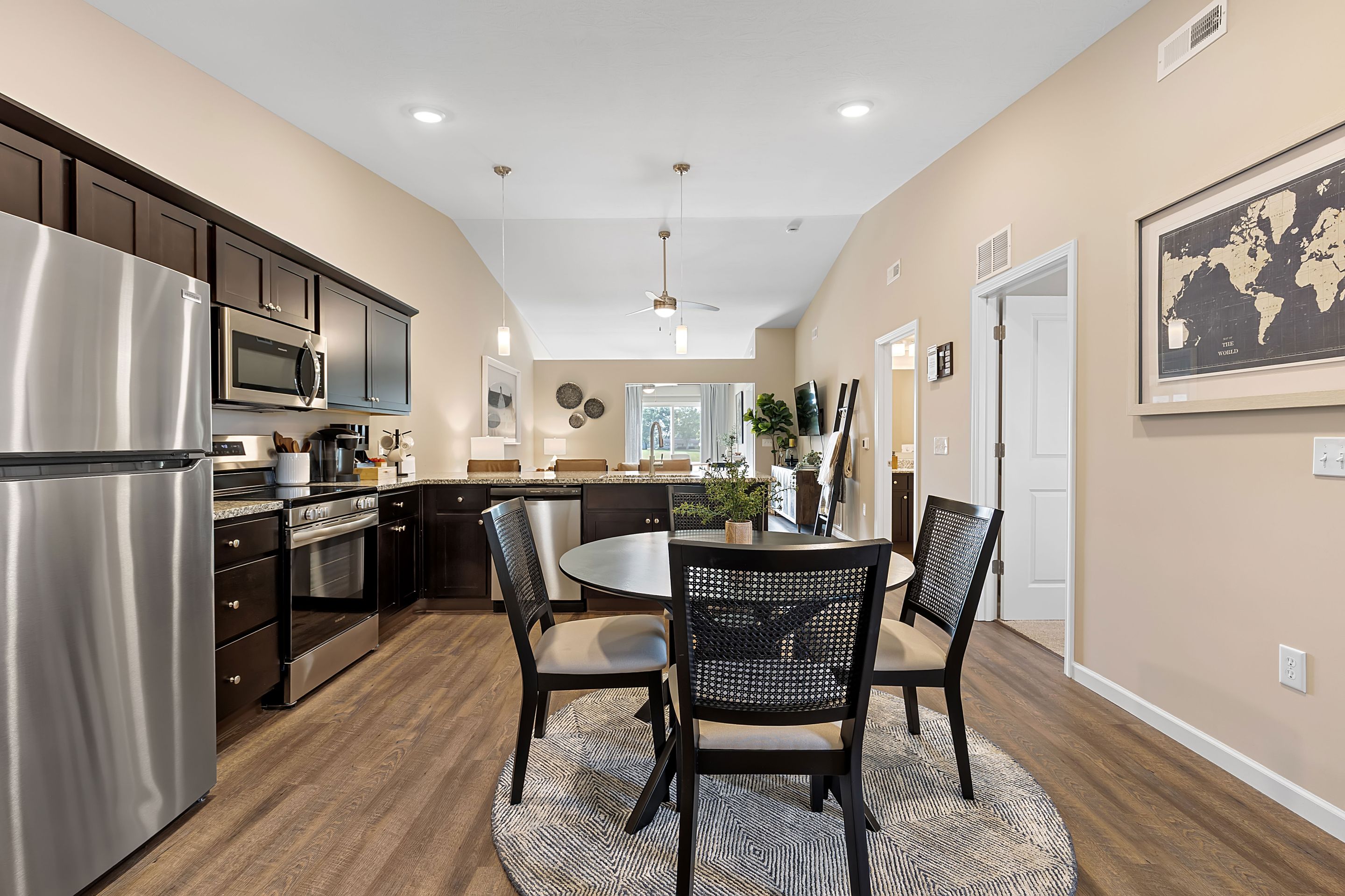 Modern kitchen and dining area with stainless steel appliances, dark cabinets, round dining table with four chairs, and a wall map, set on wood flooring.
