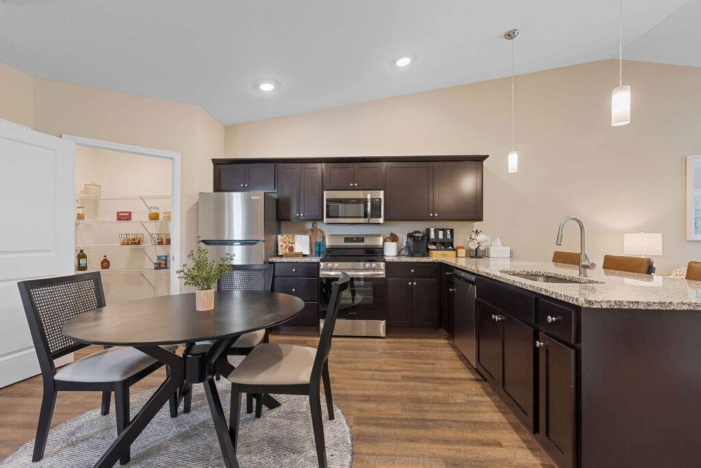 Modern kitchen with dark cabinets, stainless steel appliances, granite countertops, a round dining table with four chairs, and a pantry visible through an open door.