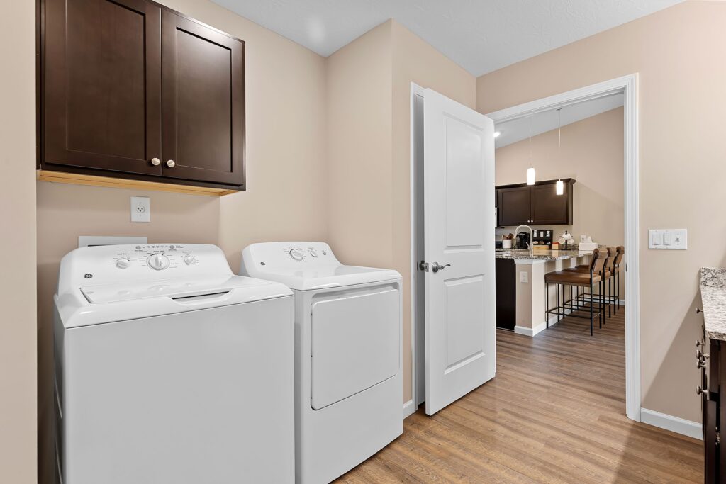 A laundry room with a white washing machine and dryer, brown wall cabinets above, wood flooring, and a view into a kitchen with barstools in the background.