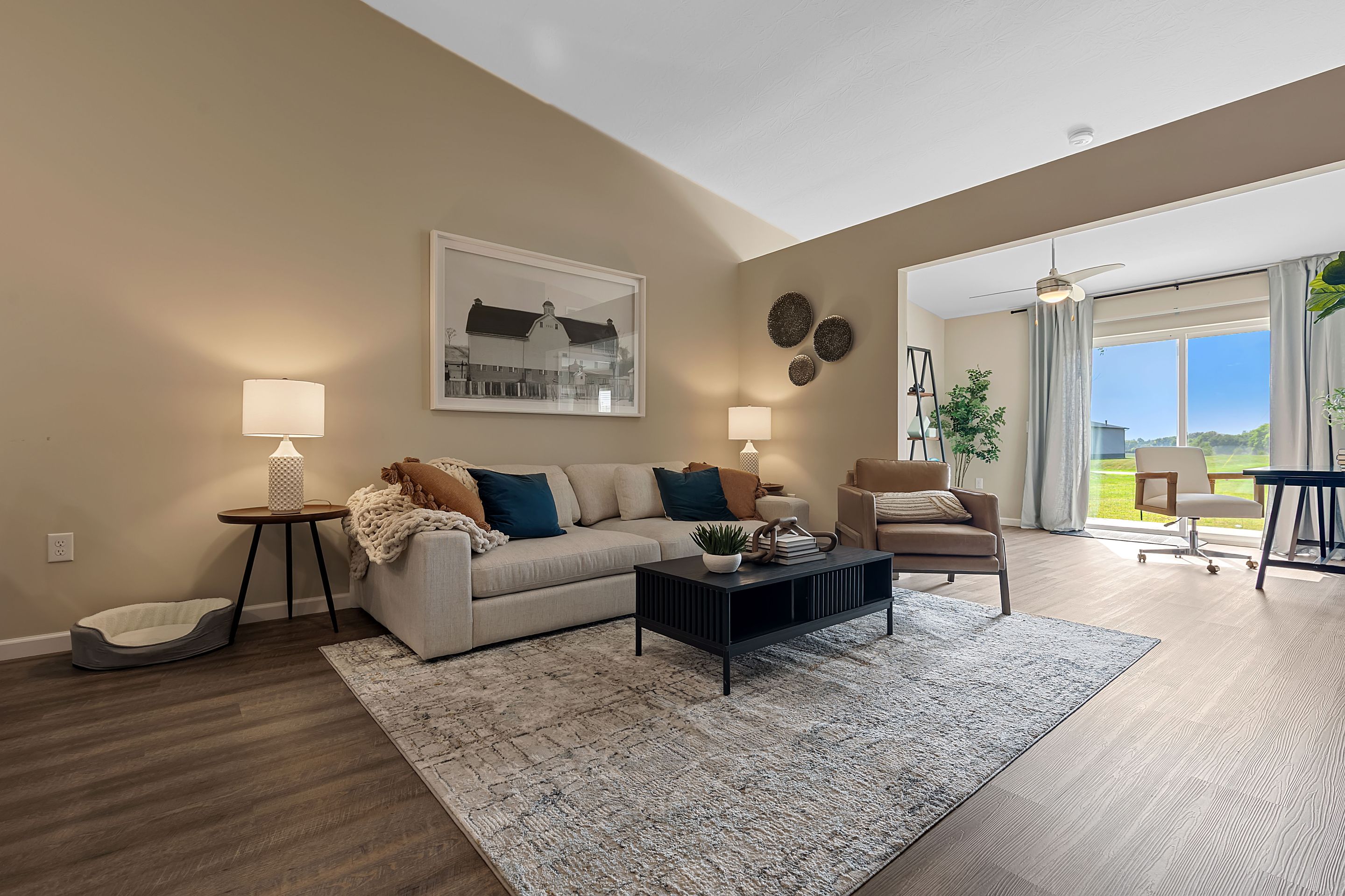 A modern living room with a beige sofa, armchair, coffee table, lamps, wall art, and large windows facing a patio with outdoor seating and green views.