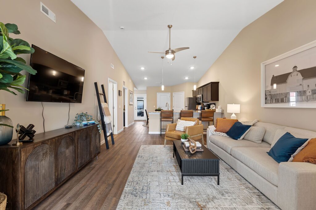 Modern living room and kitchen with neutral walls, hardwood floors, a beige sofa, wall-mounted TV, plants, and decorative accents, featuring an open floor plan and high ceiling.