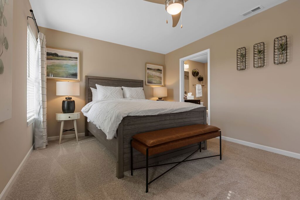 A neatly made bed with white bedding sits in a beige-carpeted bedroom, featuring a brown bench, two lamps, framed art, and a view into an adjoining bathroom.