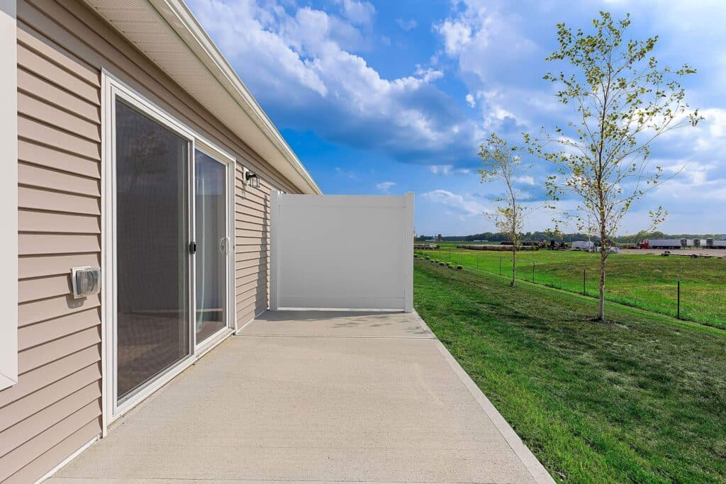 A concrete patio with sliding glass doors attached to a beige house, bordered by a white privacy fence, opens to a grassy lawn with two young trees under a partly cloudy sky.