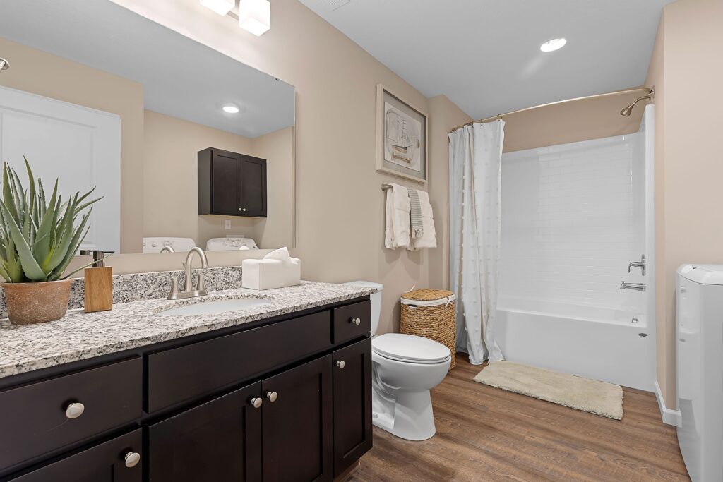 Modern bathroom with granite countertop, dark wood cabinets, a potted plant, toilet, wicker laundry basket, white shower curtain, and wood-look flooring.