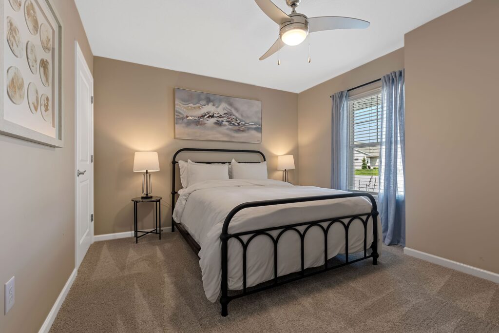 A neatly made bed with white bedding stands in a beige-walled bedroom, featuring two bedside tables with lamps, a ceiling fan, artwork, and a window with blue curtains.