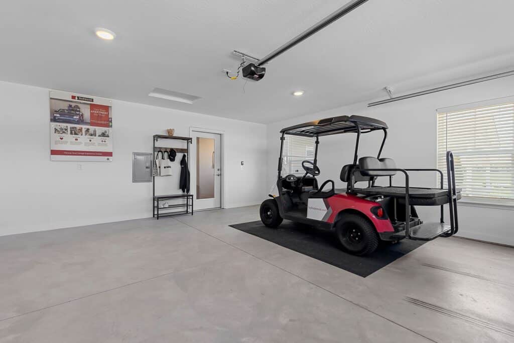 A red and black golf cart is parked on a mat in a clean, spacious garage with white walls, a window with blinds, a poster, and a coat rack.