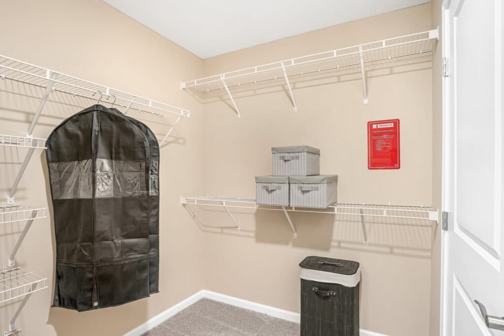 A beige walk-in closet with white wire shelves, a hanging black garment bag, three gray storage boxes, and a black hamper on carpeted flooring.