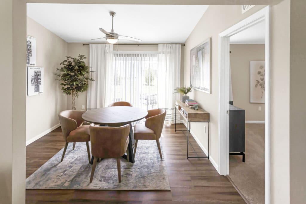 A dining room with a round table, four chairs, a console table, a potted plant, large window with sheer curtains, and hardwood floors.